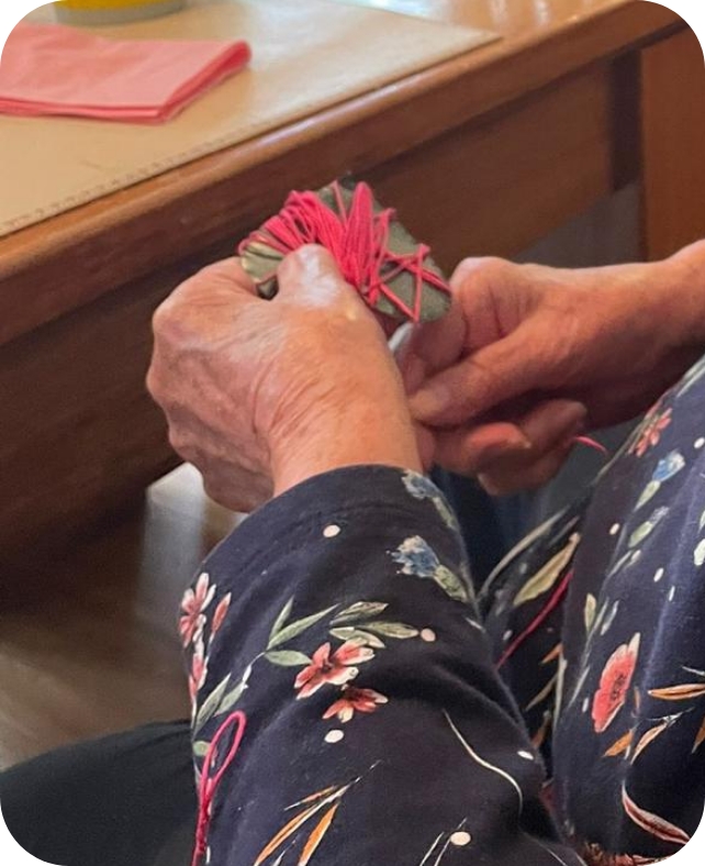 Hände einer Bewohnerin beim Umwickeln einer Bastelarbeit mit rosa Wolle